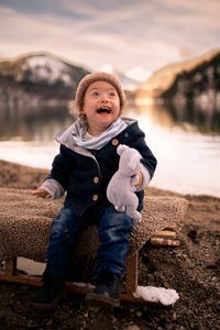 Fotoshooting mit Bild von süßem lachenden Kind das im Winter auf einem Schlitten am Alpsee, in Füssen, sitzt. Im Hintergrund sind Berge und der Alpsee gut zu erkennen. In der Hand hält das Kind ein Koala Kuscheltier.