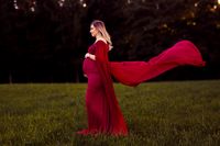 Schwangere Frau in rotem Wurfkleid steht auf einer Wiese mit Wald im Hintergrund, Fotoshooting in Kaufbeuren